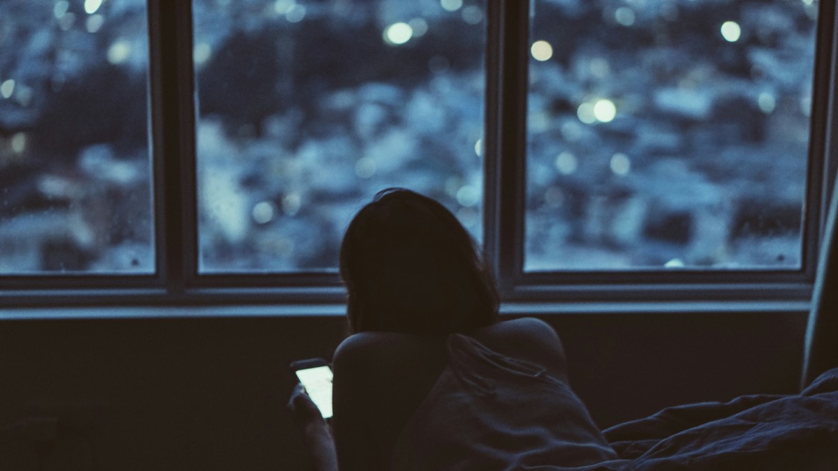 Une femme de dos regarde son téléphone devant une fenêtre, la nuit.